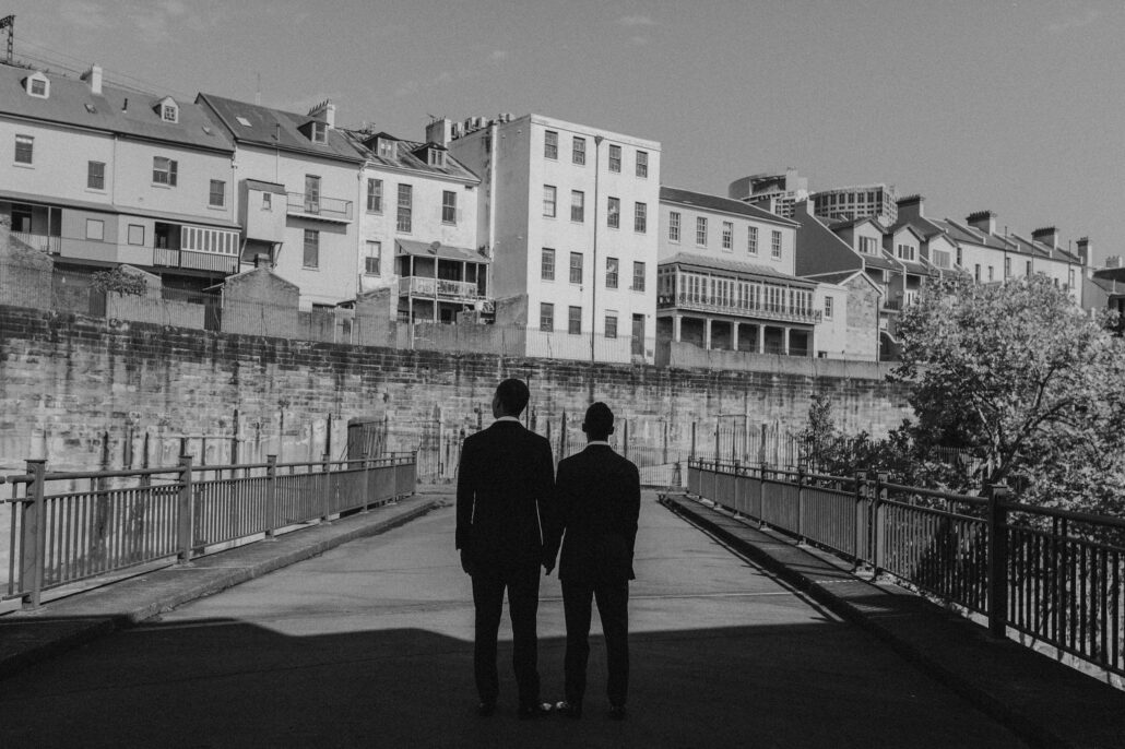 Black and white portrait of two grooms holding hands in front of old buildings
