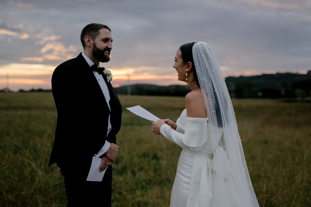Bride reading wedding vows to groom in field at sunset